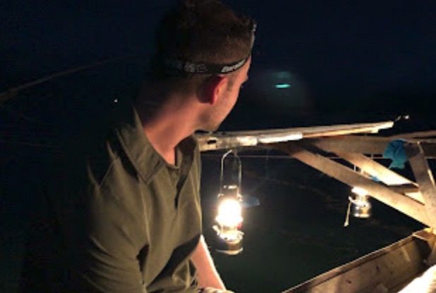 A fisherman sits beside a glowing lantern on a wooden boat on Lake Kivu after dark.